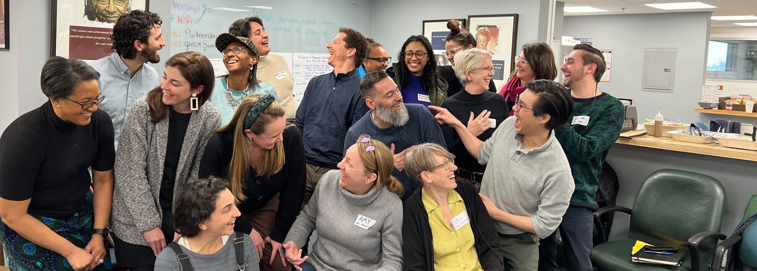 17 people gathered in an office meeting space, laughing and interacting candidly during a workshop/team-building session.