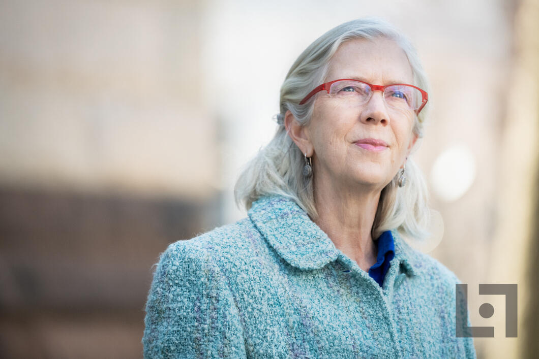 Maria Jobin-Leeds with long gray hair and red glasses standing outdoors, wearing a textured light blue coat and small dangling earrings. She looks slightly upward with a calm, thoughtful, and confident expression.